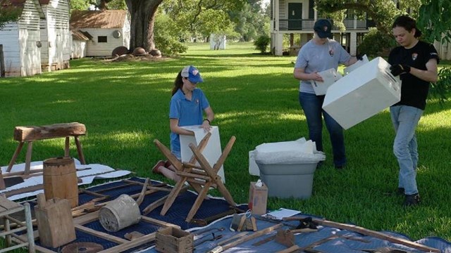 Interns working on a restoration project. Wood artifacts are laid on the floor on grass. 